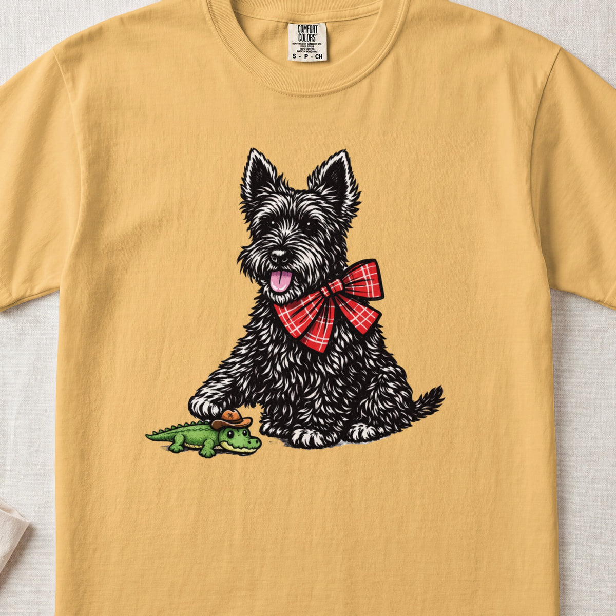 A yellow t-shirt featuring an illustration of a black Scottish Terrier wearing a red plaid bow, playing with a toy alligator.