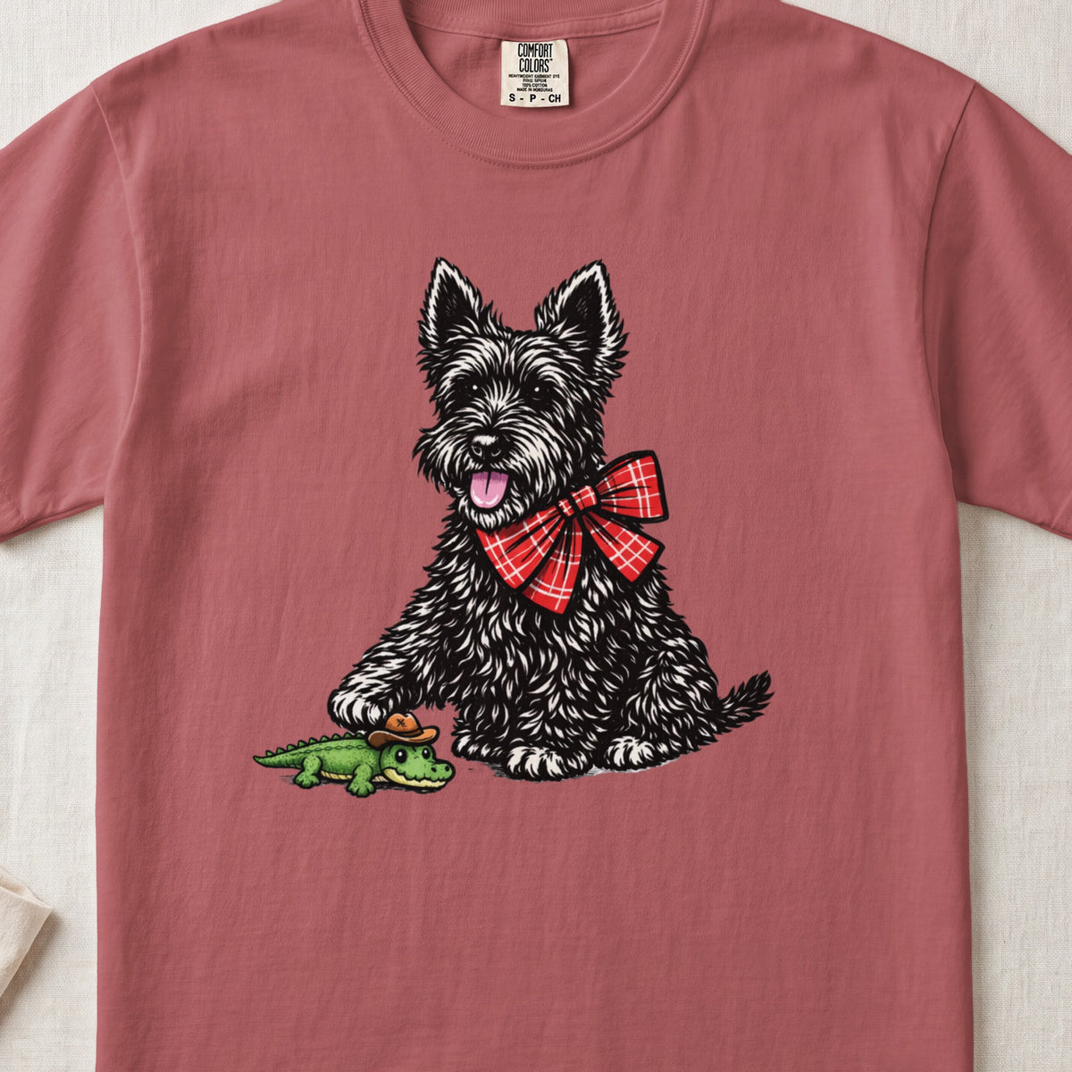A dusty rose t-shirt features a graphic of a black terrier wearing a red plaid bow, touching a tiny alligator wearing a cowboy hat.