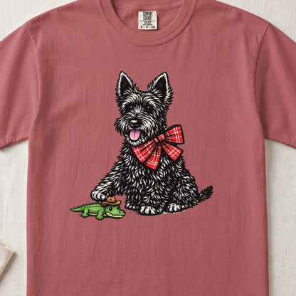 A dusty rose t-shirt features a graphic of a black terrier wearing a red plaid bow, touching a tiny alligator wearing a cowboy hat.