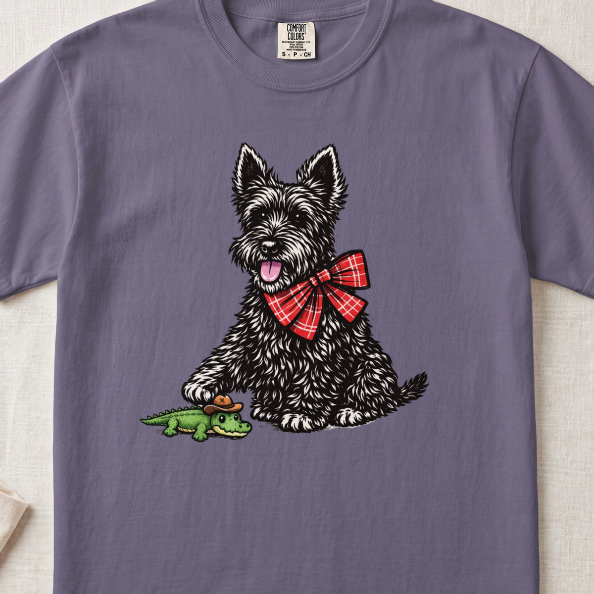 Purple t-shirt featuring an illustration of a black terrier with a red bow, playing with a toy alligator.