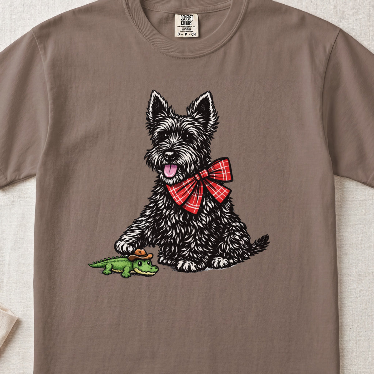 Brown crewneck t-shirt featuring a graphic of a black Scottish Terrier wearing a red plaid bow and touching a small toy alligator wearing a cowboy hat.