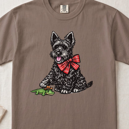 Brown crewneck t-shirt featuring a graphic of a black Scottish Terrier wearing a red plaid bow and touching a small toy alligator wearing a cowboy hat.