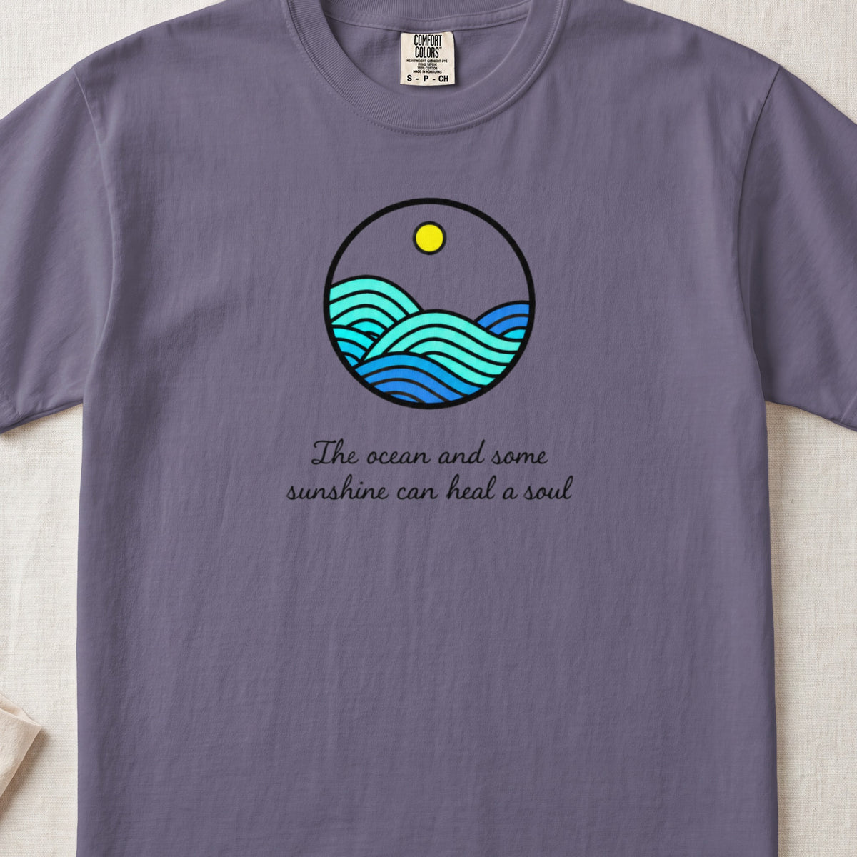 A purple t-shirt featuring a circular ocean graphic with a yellow sun and text underneath.