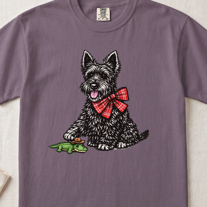 Muted purple t-shirt featuring a black terrier with a red plaid bow touching a toy alligator.