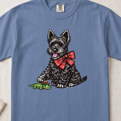 A blue t-shirt printed with a Scottish Terrier wearing a red plaid bow tie and touching a toy alligator wearing a cowboy hat.